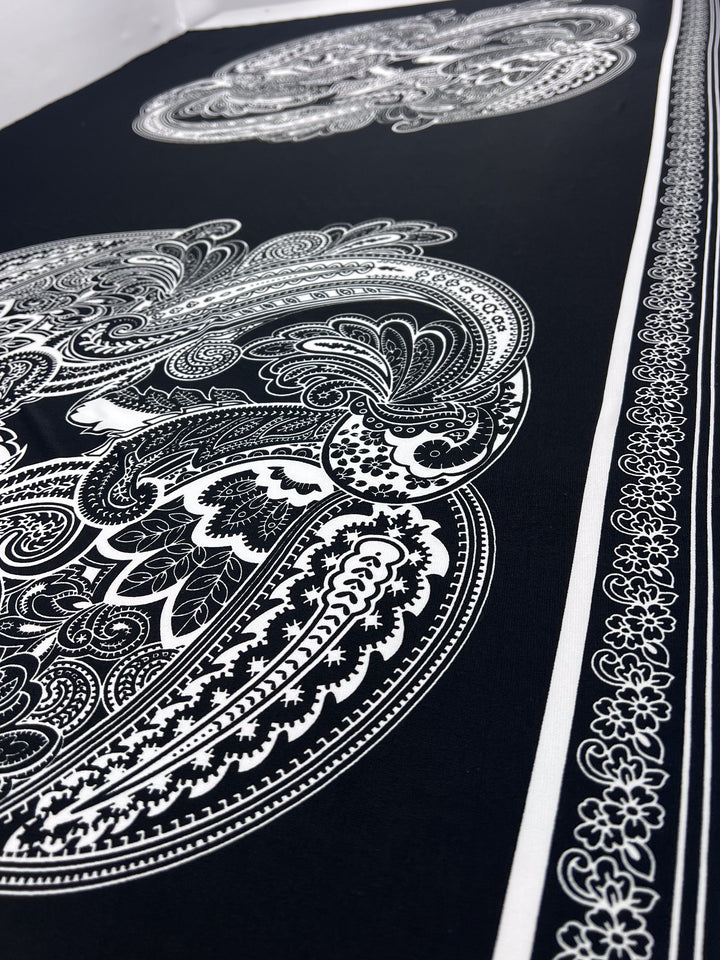 A close-up of Super Cheap Fabrics' Printed Lycra - Antoinette Paisley (85cm Panel), a black medium-weight polyester/spandex with intricate white paisley and floral patterns, detailed ovals, and decorative borders.