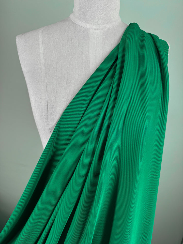 A white mannequin is adorned with Super Cheap Fabrics' Rib Knit 1x1 - Lush Meadow, a medium-weight green fabric draping diagonally over one shoulder, highlighting its textured folds against a neutral backdrop.