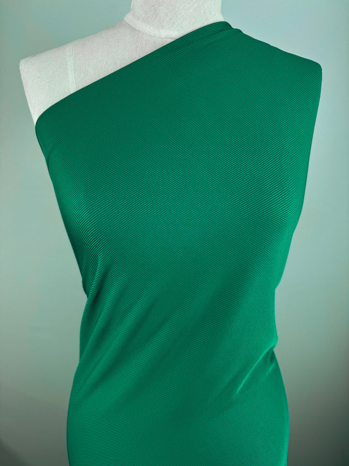 A mannequin draped in Super Cheap Fabrics' Rib Knit 1x1 - Lush Meadow - 130cm, featuring a vibrant green, medium weight rib knit fabric in a one-shoulder design against a soft, muted green background.