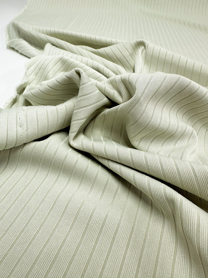 A close-up of "Rib Knit - Celadon Tint - 168cm" by Super Cheap Fabrics, this medium weight, light green knit fabric features subtle vertical stripes and a gentle texture with soft folds against a plain, light background.