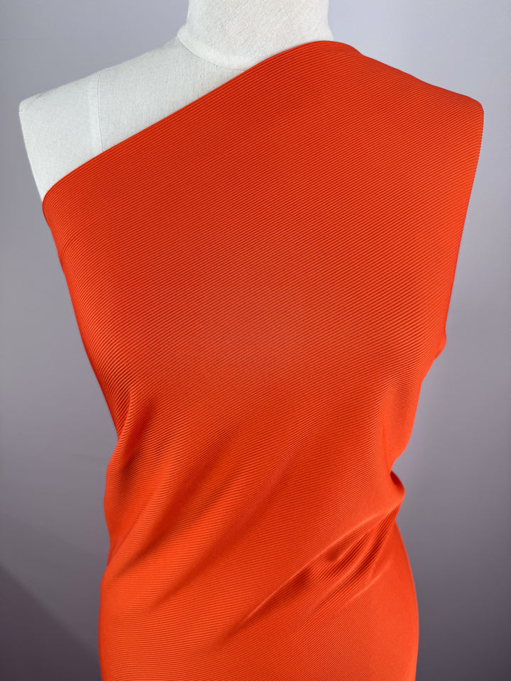 A mannequin draped with Super Cheap Fabrics' Rib Knit 1x1 - Tango (127cm) in vibrant orange, showcasing an asymmetrical one-shoulder style against a neutral gray background that highlights its unique texture.