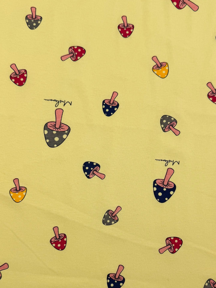The "Printed Jersey - Shroom" by Super Cheap Fabrics features a whimsical yellow design with red, yellow, and blue polka-dotted mushrooms and "Shroom" in cursive. This medium-weight, two-way stretch fabric is perfect for vibrant projects requiring charm and versatility, measuring 160cm wide.
