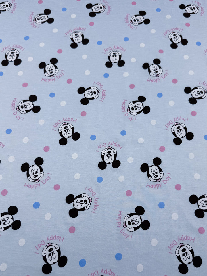 Super Cheap Fabrics' "Printed Jersey - Cheers Ears - 150cm" is a medium-weight fabric with cartoon mouse faces and smiling expressions on a light blue background, adorned with purple, pink, and blue polka dots, plus "Happy Day!" in pink script. It offers a delightful two-way stretch.