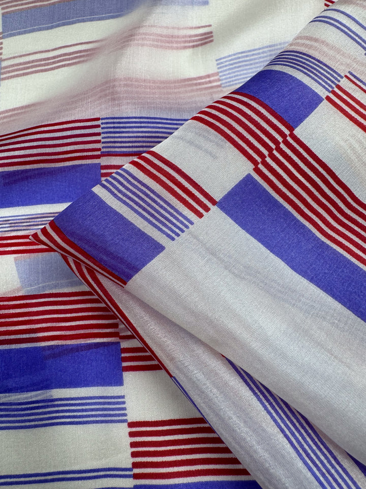The "Pure Silk - Mod Stripe - 140cm" by Super Cheap Fabrics features a dynamic geometric pattern with red and blue stripes on a white background. The varying stripe thickness and direction create a checkered look, ideal for luxury fashion, complemented by its lightweight, smooth texture.