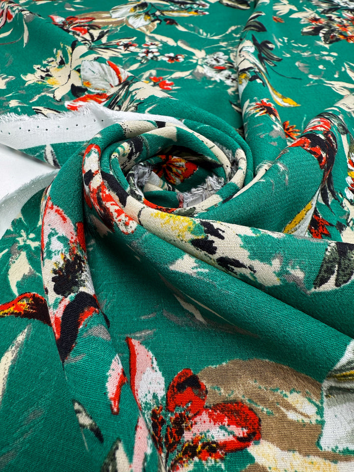 A close-up of Super Cheap Fabrics' "Printed Viscose - Tropic Bloom - 140cm" from Prato, Italy, loosely rolled to display its vibrant green background with red, white, yellow, and black floral prints.