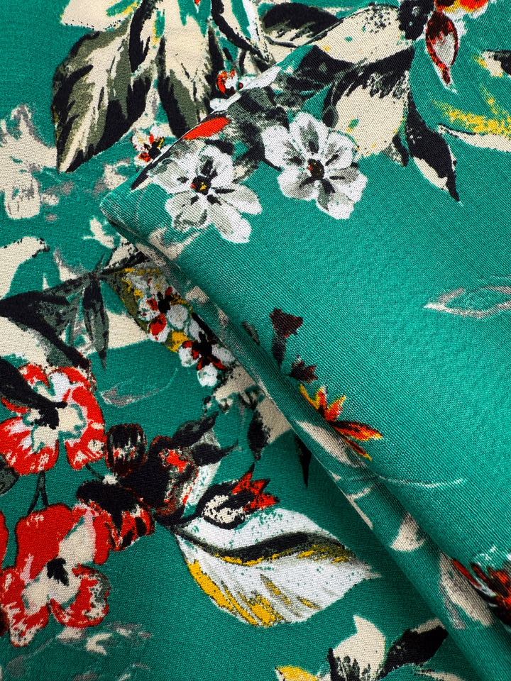 Close-up of Printed Viscose - Tropic Bloom (140cm) from Super Cheap Fabrics, featuring a vibrant green base with red, white, yellow, and black flowers plus green and cream leaves in a layered floral pattern—sourced from Prato, Italy.