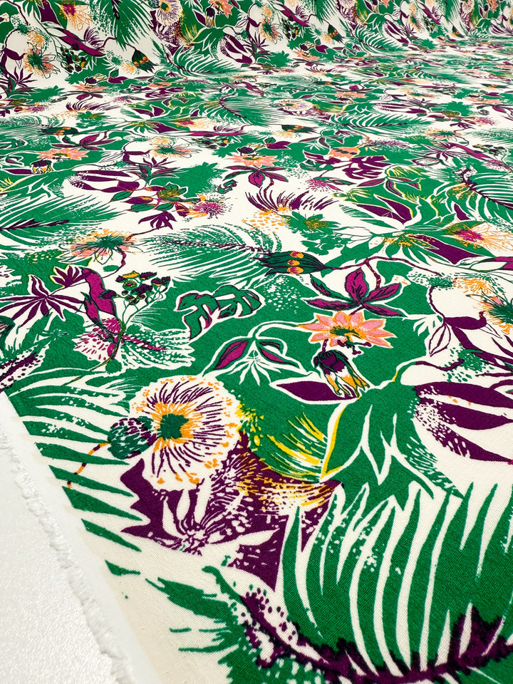 A close-up of Super Cheap Fabrics' Printed Viscose - Jungle Orchid (142cm), featuring a vibrant tropical floral pattern in green, purple, yellow, and orange, shown flat with the fabric edge visible on the left.