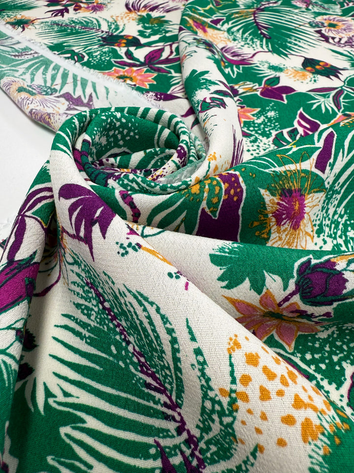A close-up of Super Cheap Fabrics' Printed Viscose - Jungle Orchid (142cm), featuring a vibrant green, purple, and yellow tropical floral and leaf design on white, artistically folded and rolled into a spiral. Deadstock from Prato, Italy.