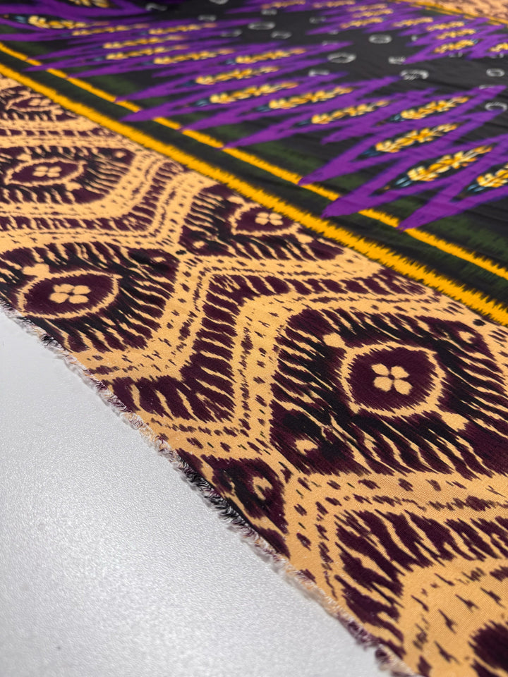 A close-up of Super Cheap Fabrics’ Printed Viscose - Regal Ikat (145cm) shows its vibrant yellow, purple, black, and beige geometric patterns with visible texture and frayed edges on a white surface.