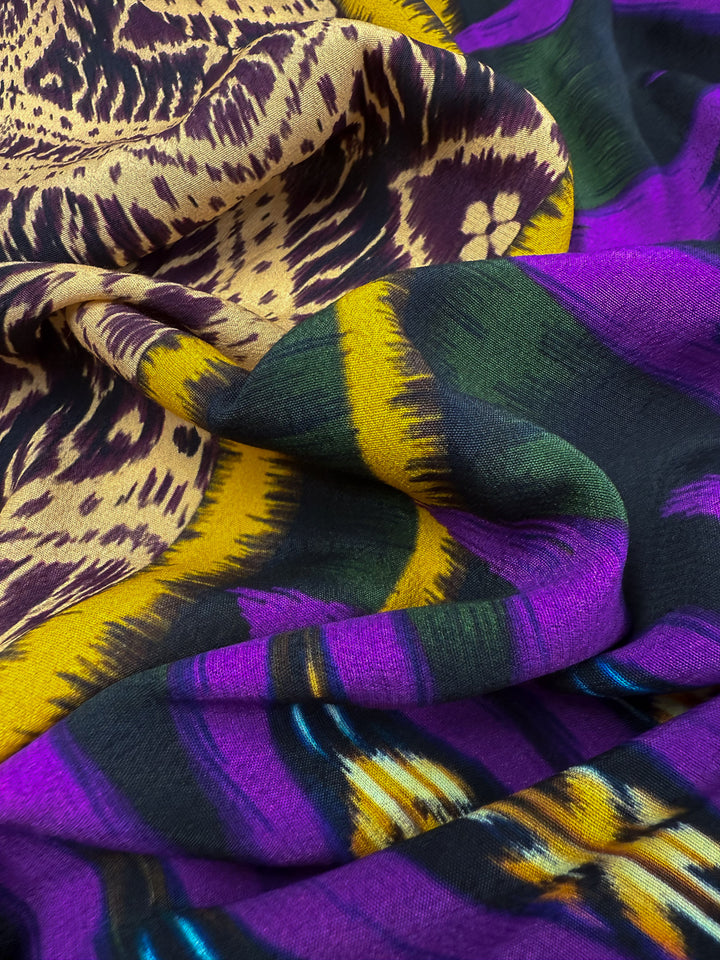 A close-up of Super Cheap Fabrics' Printed Viscose - Regal Ikat (145cm), showcasing vibrant yellow, purple, green, and black abstract swirls with a slightly crumpled texture.