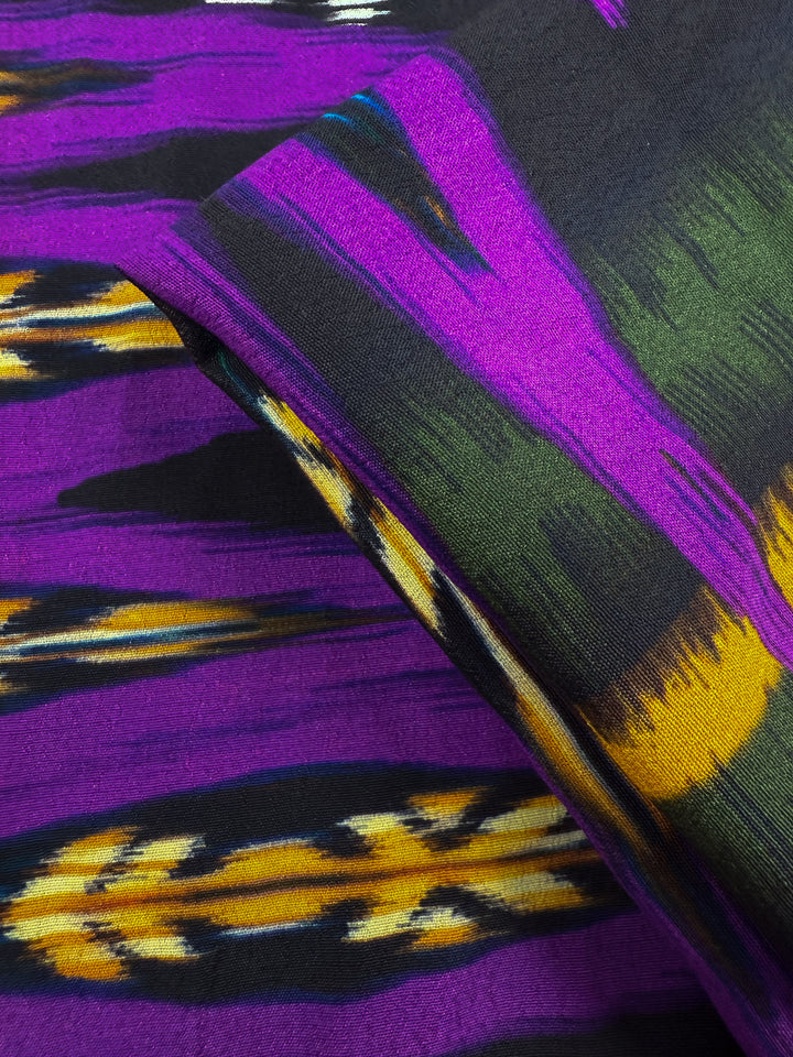A close-up of Super Cheap Fabrics' Printed Viscose - Regal Ikat (145cm), featuring bold purple, yellow, black, and green geometric and zigzag patterns that highlight the fabric's vibrant texture and layered design.