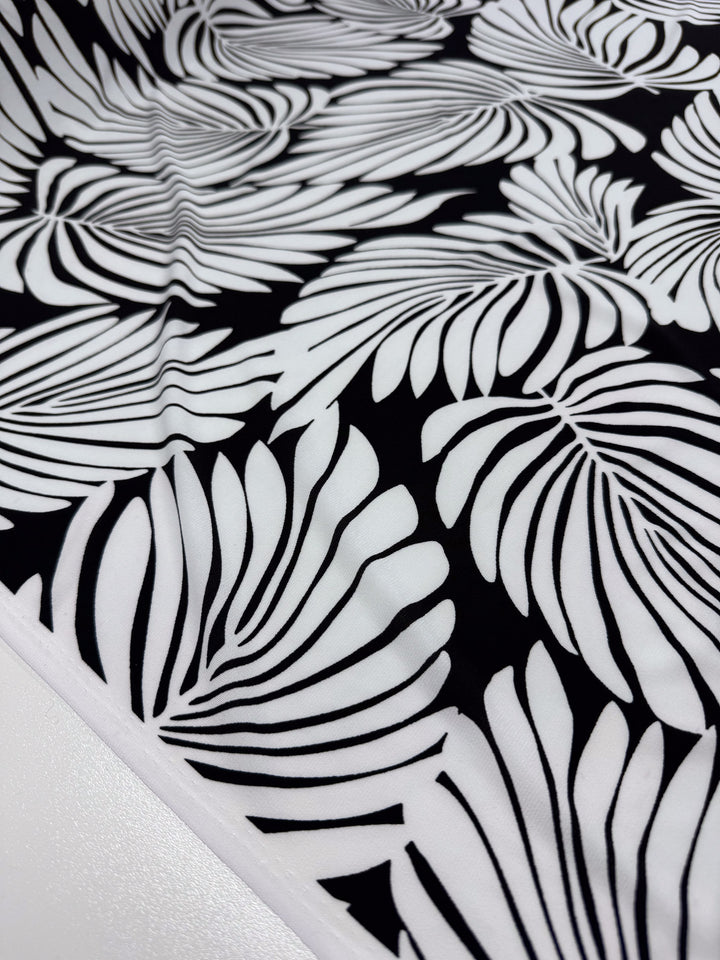 Close-up of "Printed Lycra - Palma" by Super Cheap Fabrics, a medium-weight 150cm Polyester/Spandex blend with a bold black and white pattern featuring large stylized fern or palm leaves overlapping for an intricate, modern look that emphasizes contrast and texture.