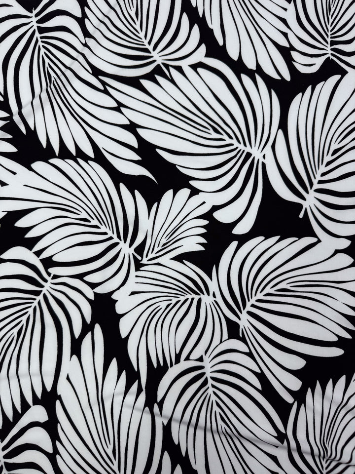 The Printed Lycra - Palma from Super Cheap Fabrics features overlapping stylized palm leaves in black and white, creating a striking contrast and dynamic design on this medium-weight fabric. Size: 150cm.