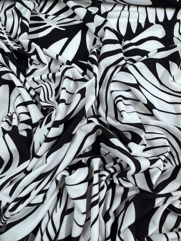 Super Cheap Fabrics' Printed Lycra - Palma (150cm) showcases a bold, black and white abstract leaf pattern with contrasting stripes and curves, creating a striking visual texture on this medium-weight fabric.