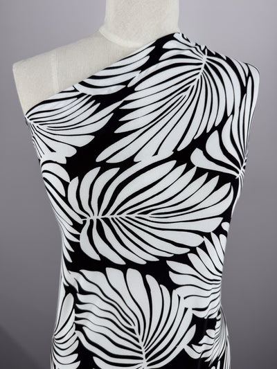 A mannequin displays the "Printed Lycra - Palma - 150cm" by Super Cheap Fabrics, a one-shoulder garment made from medium-weight Polyester/Spandex. It features bold black and white stylized leaf patterns on a soft gray background.