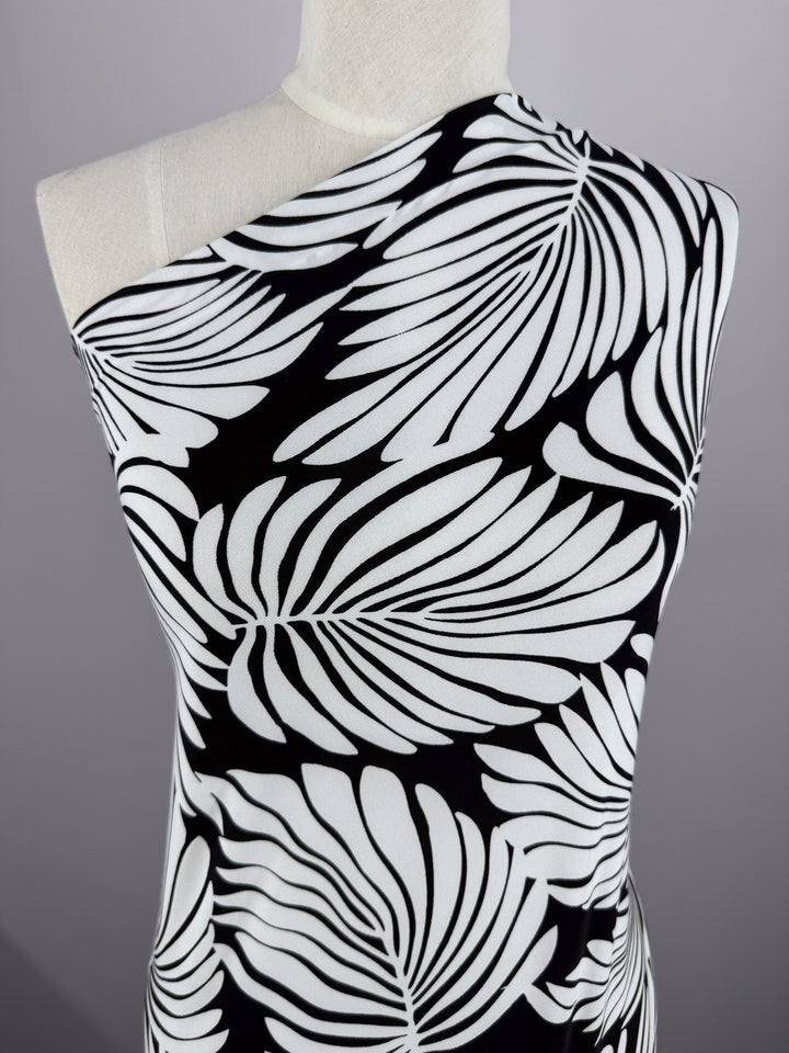 A mannequin displays the "Printed Lycra - Palma - 150cm" by Super Cheap Fabrics, a one-shoulder garment made from medium-weight Polyester/Spandex. It features bold black and white stylized leaf patterns on a soft gray background.
