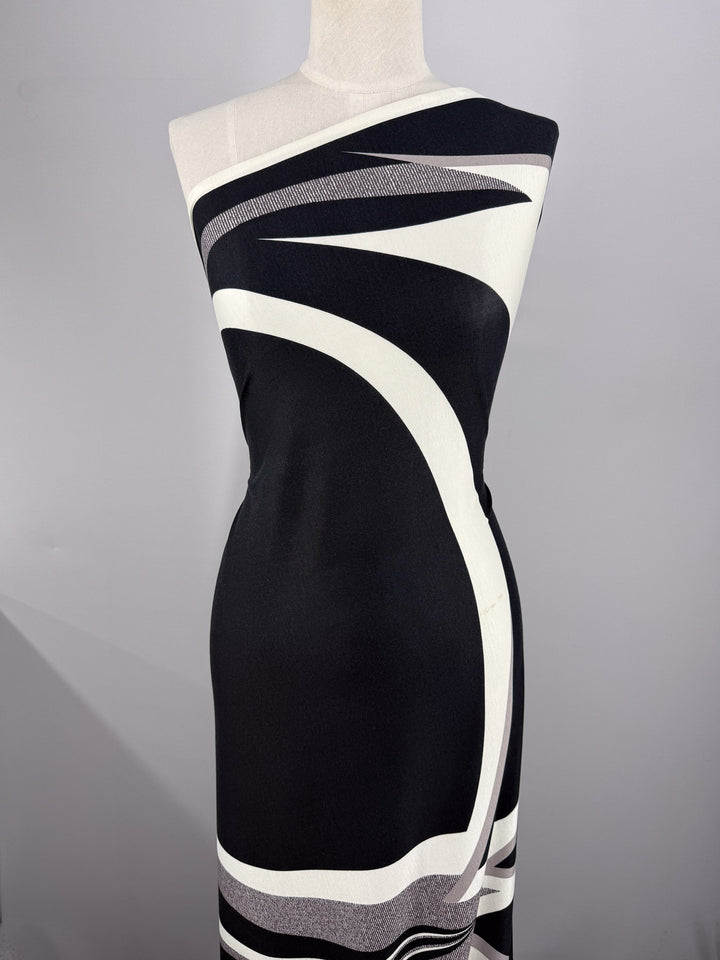 A mannequin showcases a sleeveless, asymmetrical dress made from Super Cheap Fabrics’ Viscose Knit - Curve - 145cm, featuring bold white and gray curved lines in a modern abstract pattern on breathable black fabric.