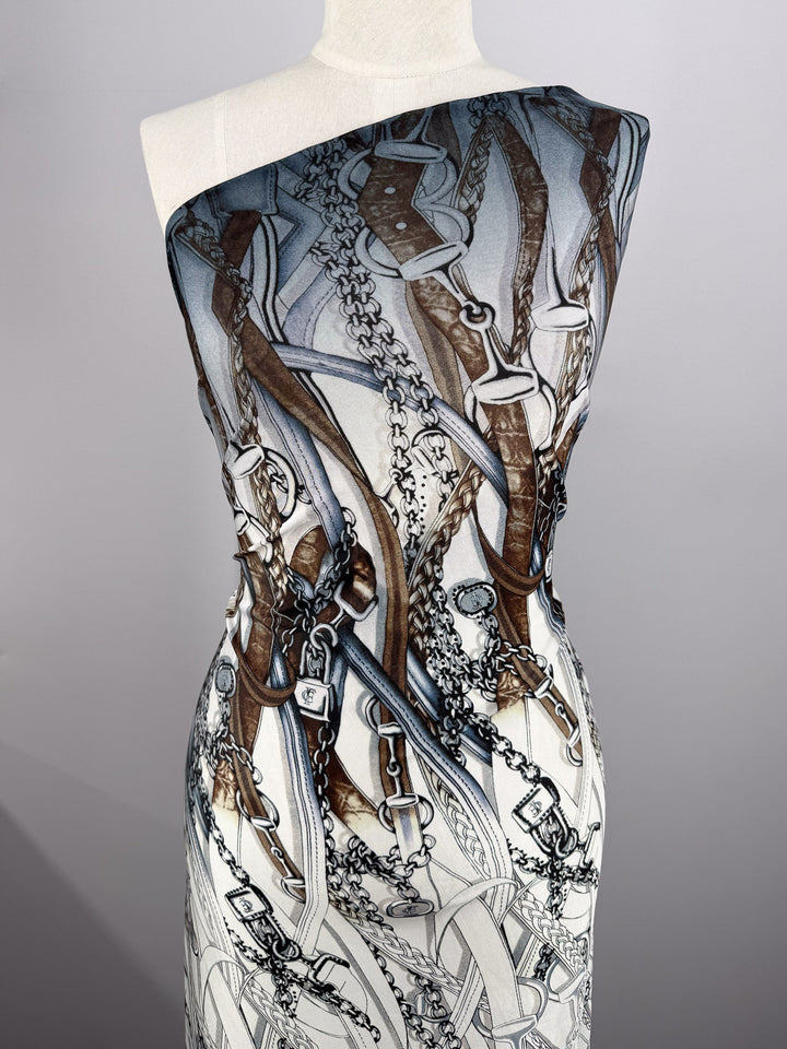 A mannequin displays Super Cheap Fabrics' Viscose Knit - Link (138cm), a stretch fabric with an elegant pattern of brown leather straps, silver chains, and buckles over a white and gray background.