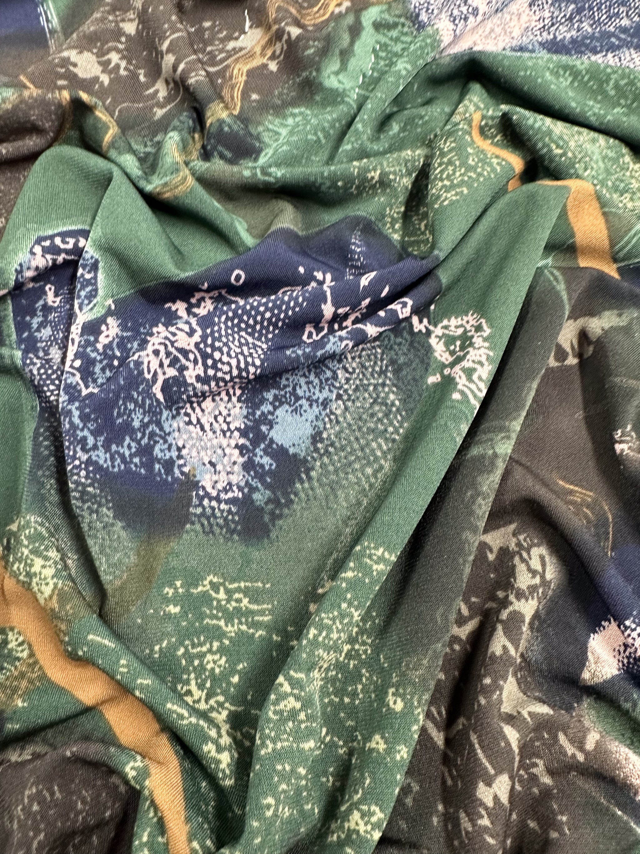 Close-up image of a crumpled, richly colored medium-weight fabric with various textures and patterns in shades of green, blue, and hints of yellow and white. The Super Cheap Fabrics Printed Lycra - Drifter - 150cm fabric appears soft and has an intricate, abstract design.