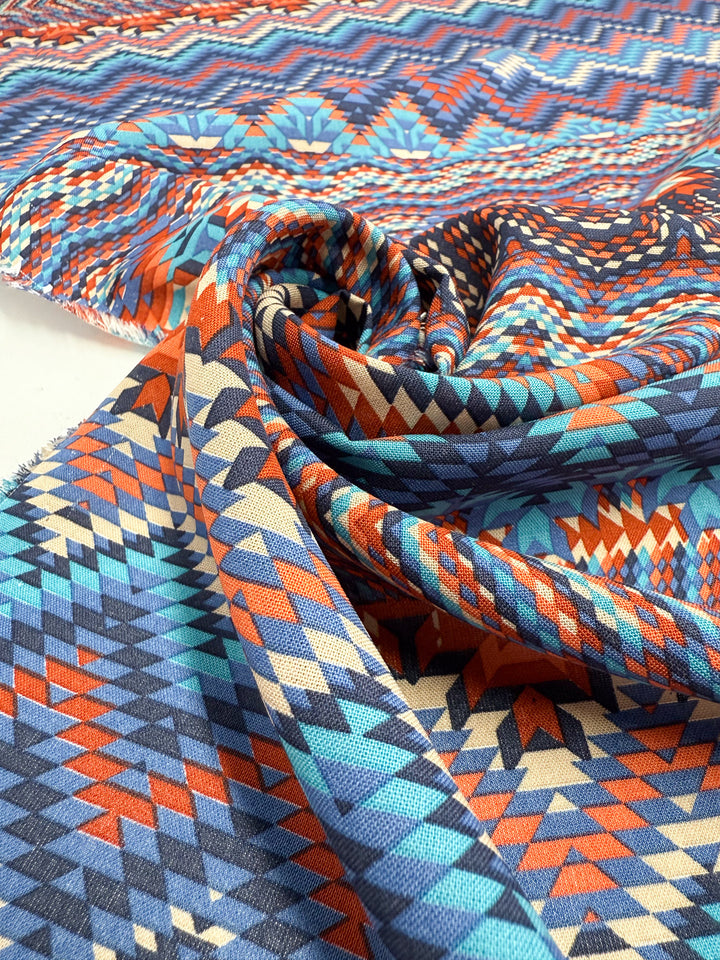 Printed Viscose - Zulu - 150cm by Super Cheap Fabrics features a vibrant geometric pattern in blue, orange, red, and beige. The soft woven fabric is displayed in gentle folds to highlight its color and texture.