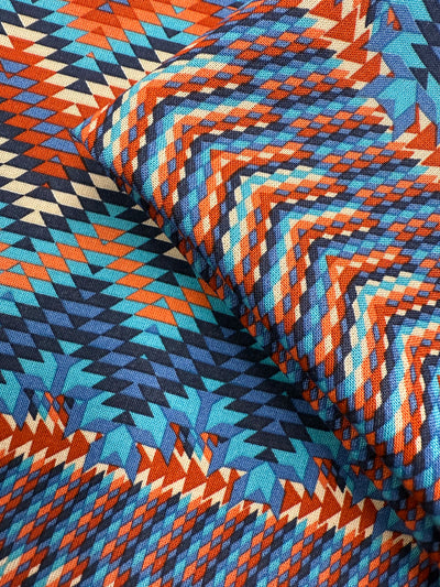 A close-up of Super Cheap Fabrics’ Printed Viscose - Zulu - 150cm shows geometric zigzag and diamond patterns in blue, orange, red, yellow, and beige from Prato, Italy. Slight folding adds depth and texture.