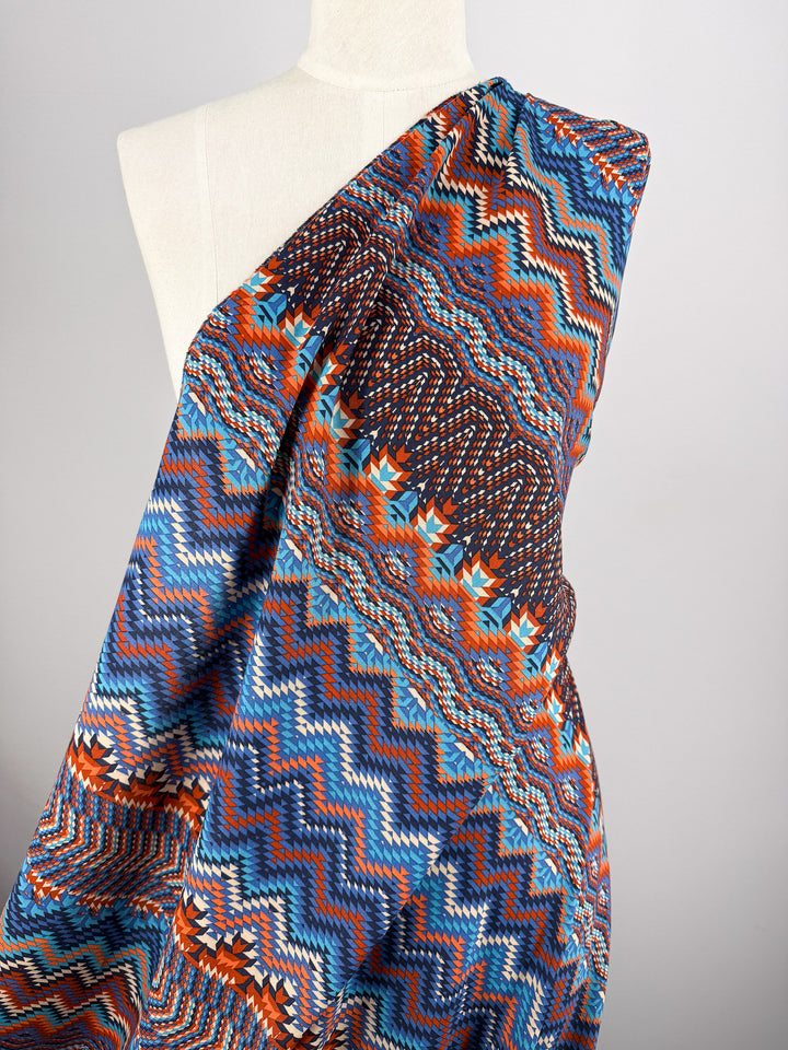 A mannequin showcases Super Cheap Fabrics' Printed Viscose - Zulu - 150cm, featuring a bold zigzag pattern in blue, orange, white, and brown, draped over one shoulder against a plain light background.