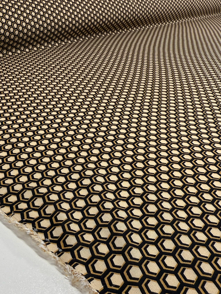 A close-up of Super Cheap Fabrics' Printed Viscose - Hive - 145cm shows lightweight fabric with black hexagons outlined in gold on beige, creating an optical illusion effect.