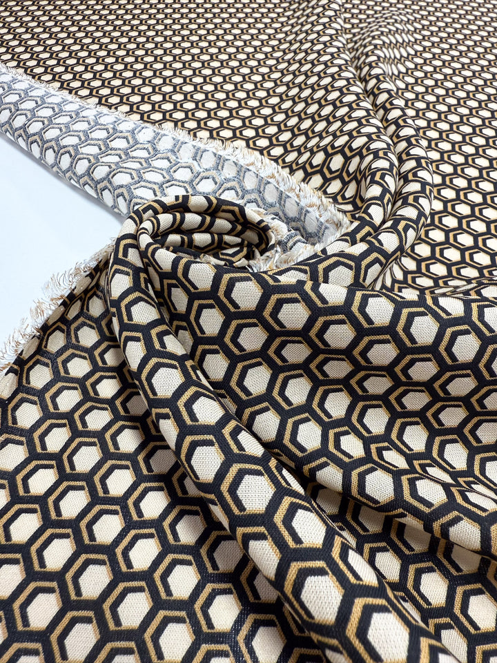A close-up of Super Cheap Fabrics' Printed Viscose - Hive (145cm) shows its lightweight Italian material with a black and gold geometric hexagon pattern on beige, draped to reveal both front and back sides.