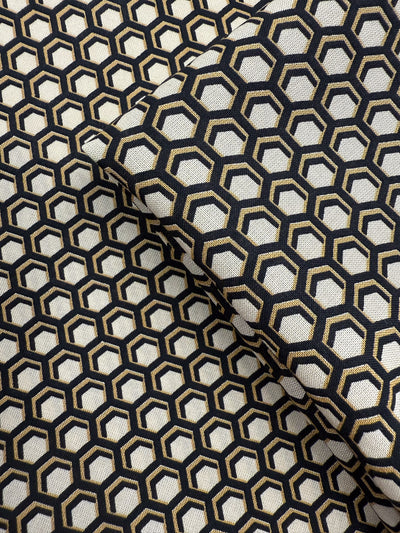 A close-up of Super Cheap Fabrics’ Printed Viscose - Hive - 145cm shows folded fabric with a black and gold geometric hexagon print on an off-white base, creating an optical illusion—ideal for lightweight projects.