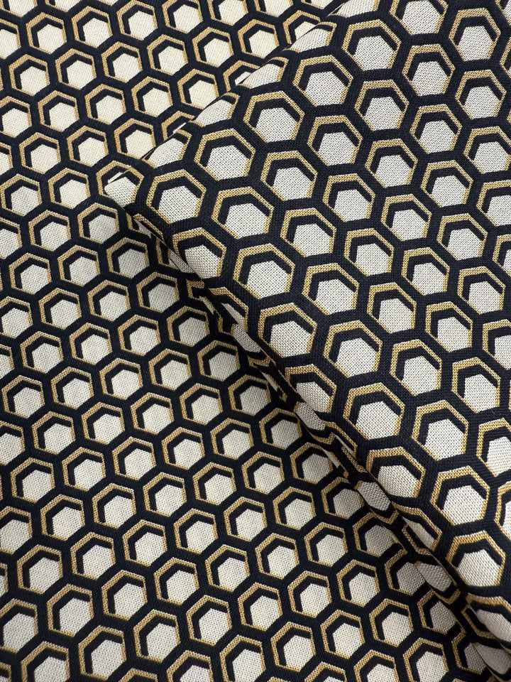 A close-up of Super Cheap Fabrics’ Printed Viscose - Hive - 145cm shows folded fabric with a black and gold geometric hexagon print on an off-white base, creating an optical illusion—ideal for lightweight projects.