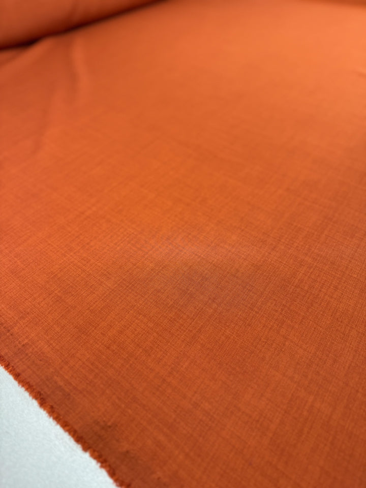 A close-up of Super Cheap Fabrics' Twill Suiting in Rust (150cm) reveals smooth orange polyester with a subtle diagonal weave and a slightly frayed left edge.
