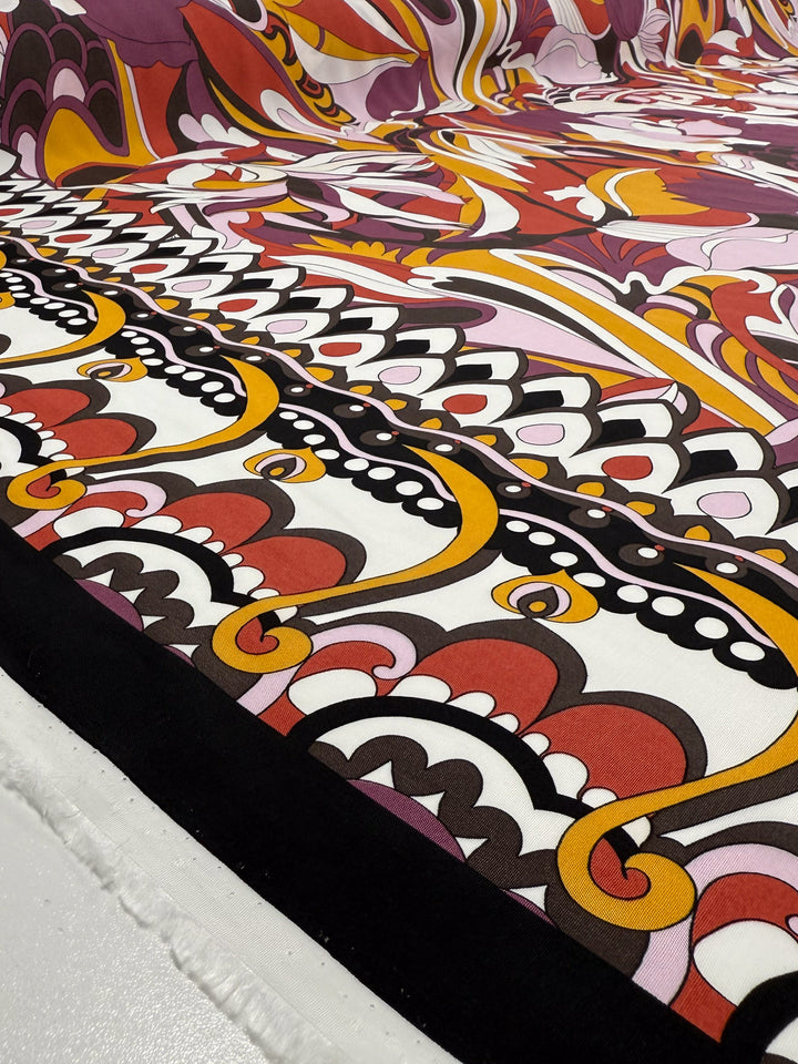 A close-up of Super Cheap Fabrics' Printed Viscose - Fantasia (145cm), featuring bold abstract red, orange, purple, yellow, black, and white patterns with swirling curved lines and organic shapes, laid flat on a white surface.