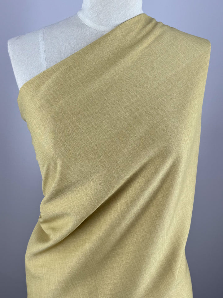 A mannequin is draped in "Twill Suiting - Cocoon - 150cm" by Super Cheap Fabrics, showcasing its light yellow, fine diagonal weave. The versatile fabric runs diagonally across the torso, exposing one shoulder against a plain backdrop.