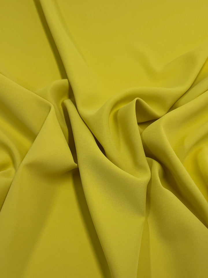 A close-up of Super Cheap Fabrics' Microfibre Suiting - Mango Mint (150cm) shows bright yellow fabric with soft folds and gentle draping, giving it a smooth texture with a slightly wrinkled look.