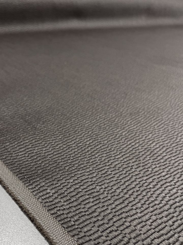 Close-up of Super Cheap Fabrics' Upholstery Jacquard - Oscar Putty (148cm), showing its textured dark gray surface, fine ridges, subtle pattern, heavy weight feel, and fabric border along the edge.