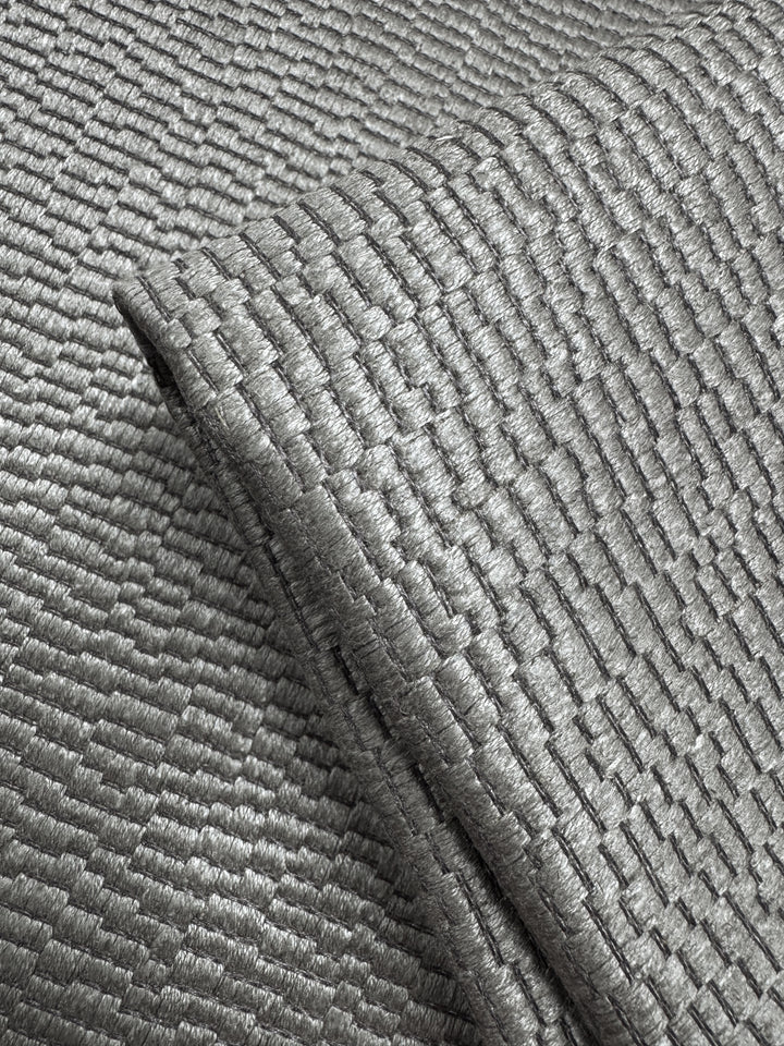 Close-up of Super Cheap Fabrics' Upholstery Jacquard - Oscar Putty (148cm): a shiny, silver-gray heavyweight fabric with a geometric basket-weave pattern that reflects light, showcasing its intricate weave and metallic look.