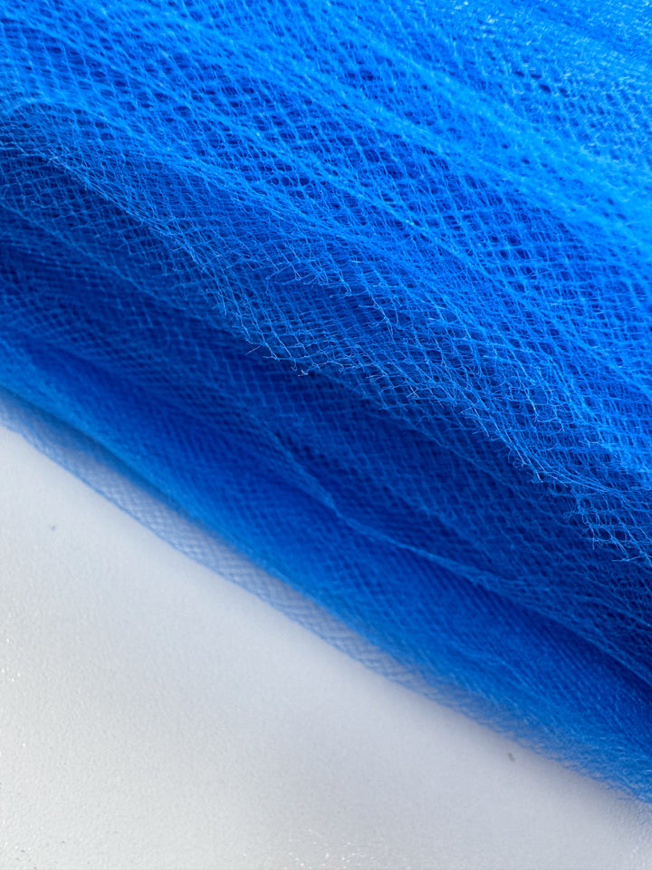 A close-up of Super Cheap Fabrics’ Soft Tulle - Royal - 150cm shows its fine net texture in bright blue, resting on a white surface—ideal for bridal veils or crafts.