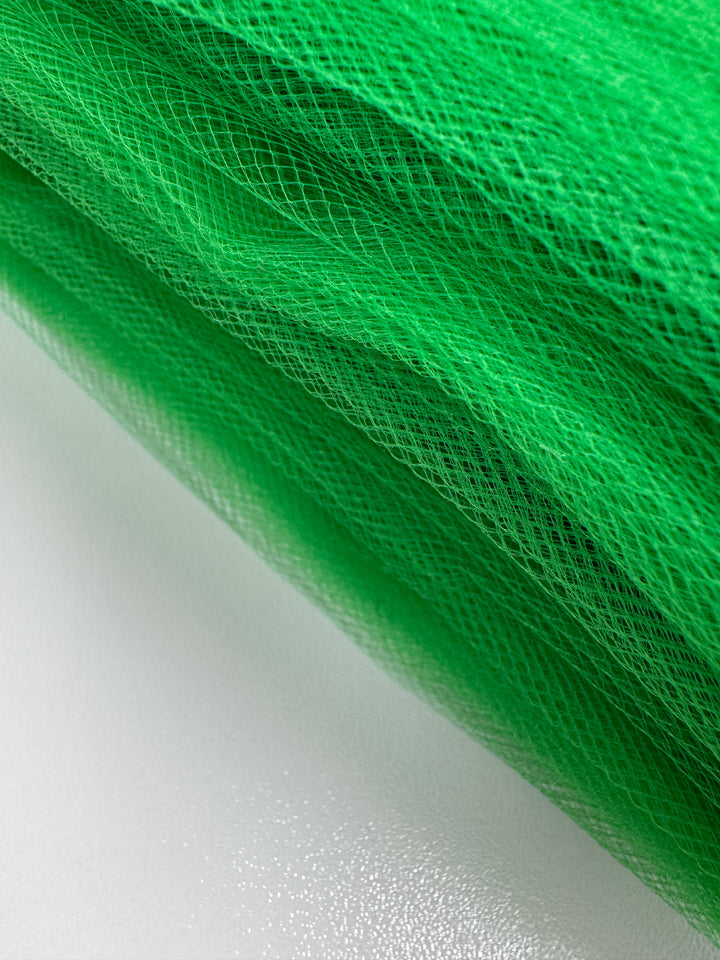 A close-up of Super Cheap Fabrics' Soft Tulle - Emerald - 150cm, a lightweight and versatile fabric, rolled on a white surface to showcase its fine mesh texture and layered look.
