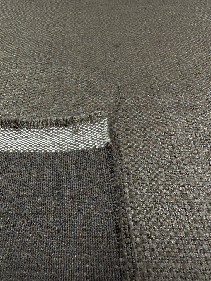 A close-up of Super Cheap Fabrics' Upholstery Basket Weave - Dorian Putty (145cm) reveals its textured, heavy-weight woven pattern. A folded corner shows a white stripe underneath, while frayed edges lend a rustic touch to this upholstery piece.