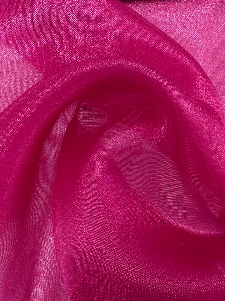 A close-up of Super Cheap Fabrics' Organza - Very Berry (150cm), showing sheer, shimmery pink fabric draped in soft folds that create a wavy, textured pattern with elegant highlights and shadows.