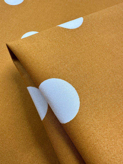 A close-up of the Cotton Sateen - Polka Yam fabric by Super Cheap Fabrics shows a mustard yellow base with large white polka dots. This 125cm wide cotton sateen fabric has a smooth texture and subtle sheen, making it ideal for creating playful, modern dresses.