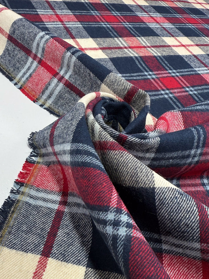 A close-up of Super Cheap Fabrics' Flannel Tartan - Highland (145cm), a medium weight cotton in red, navy, cream, and gray Scottish-style plaid, swirled to reveal its texture and frayed edge on a white surface.