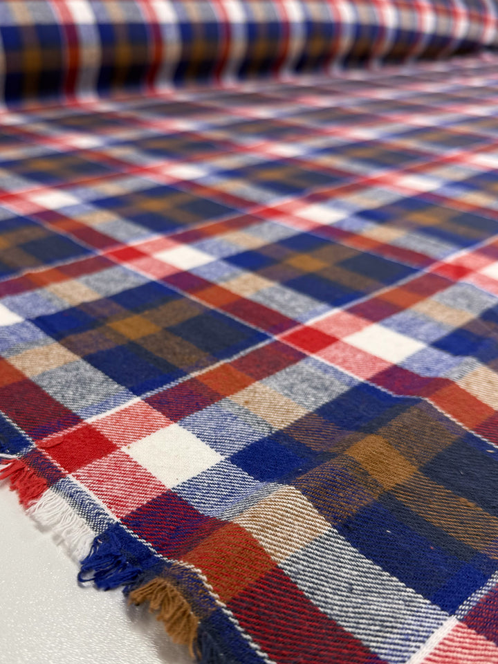 A close-up of Super Cheap Fabrics' Flannel Plaid - Autumn Rust Check (145cm) shows red, blue, white, and brown stripes on a smooth medium weight fabric with slightly frayed edges.