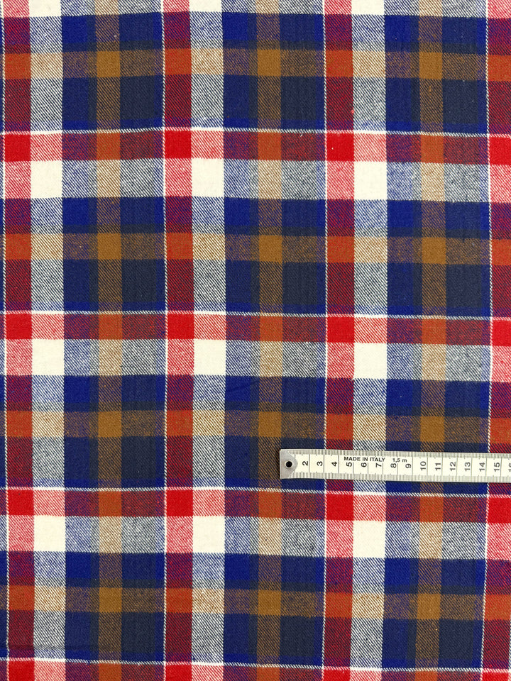 A close-up of Super Cheap Fabrics’ Flannel Plaid - Autumn Rust Check (145cm) shows the red, blue, white, and mustard pattern with a ruler at the bottom right displaying centimeter scale.