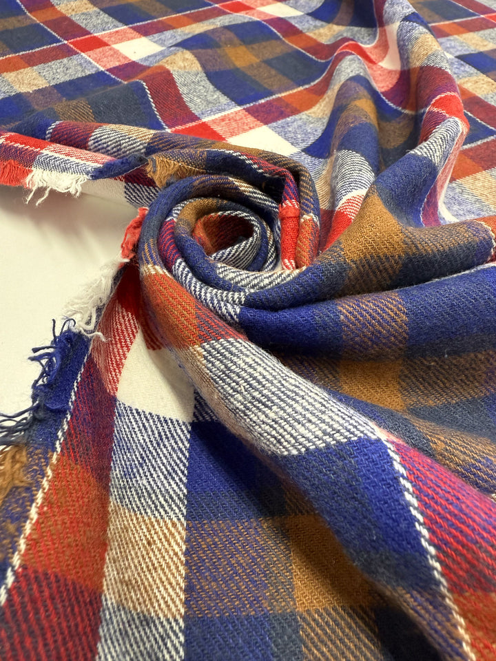 A close-up of Super Cheap Fabrics’ Flannel Plaid - Autumn Rust Check (145cm), showcasing colorful red, blue, mustard yellow, and white stripes with frayed edges on a white surface.