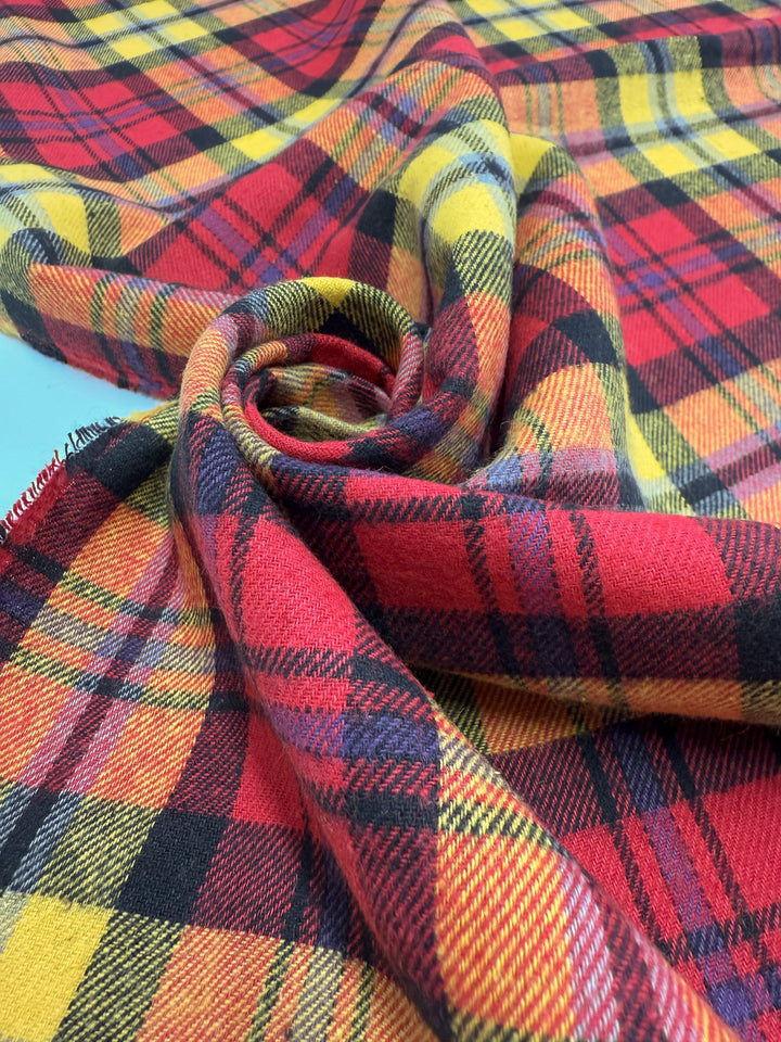 A close-up of Super Cheap Fabrics' Flannel Tartan - Edinburgh Flame (145cm), featuring a bold red, yellow, and black plaid pattern, artfully twisted into a spiral to evoke a striking punk-inspired style.