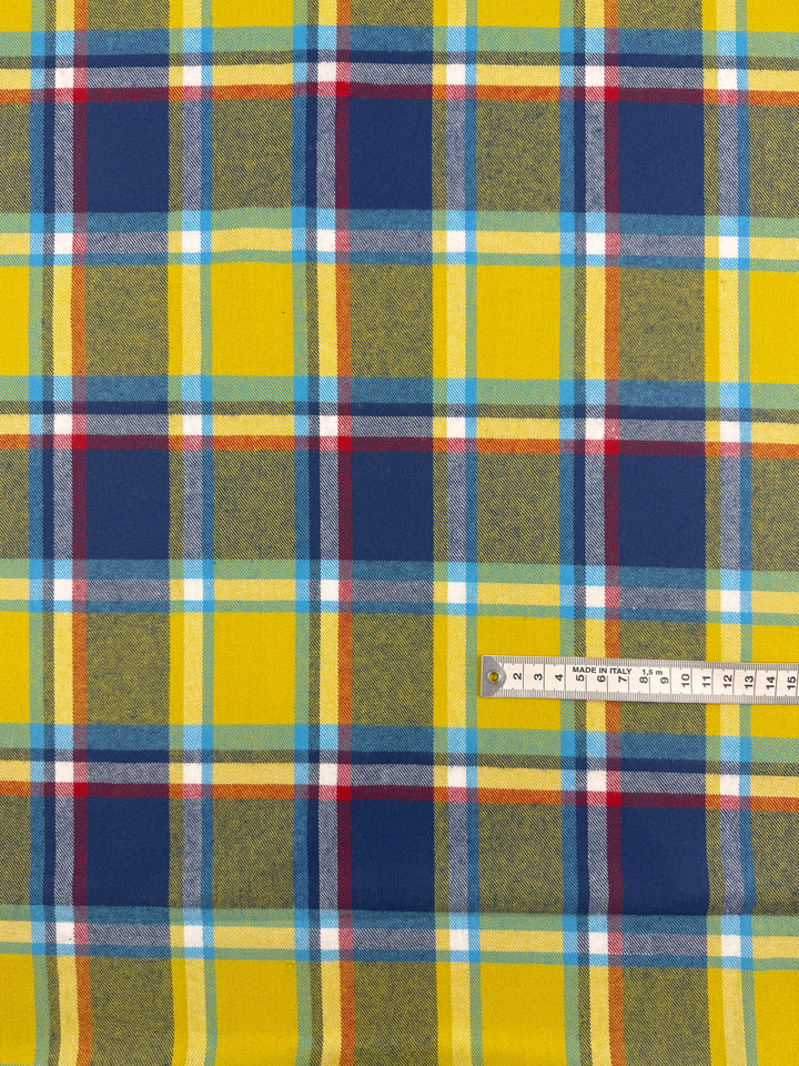 The Flannel Plaid - Sunshine Heritage (145cm) by Super Cheap Fabrics features a colourful pattern with yellow, blue, red, and white intersecting lines of varying thicknesses. A measuring tape on the right shows the fabric’s scale.