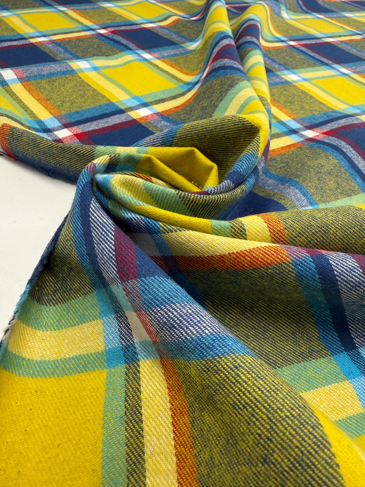 A close-up of Super Cheap Fabrics' Flannel Plaid - Sunshine Heritage (145cm) showcases its yellow, blue, red, and white checks and soft texture draped on a flat surface.