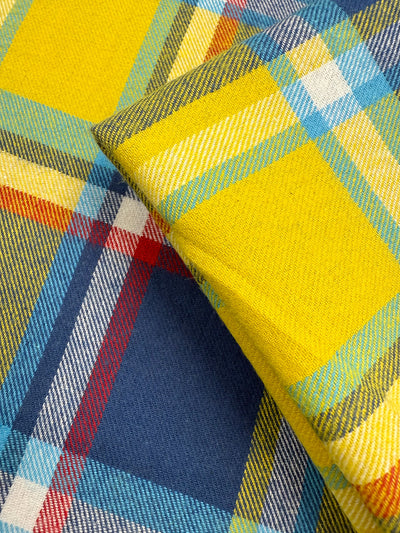 Close-up of Super Cheap Fabrics' Flannel Plaid - Sunshine Heritage (145cm), showing two overlapping pieces with a vibrant plaid pattern in bright yellow, blue, red, white, and green intersecting stripes.