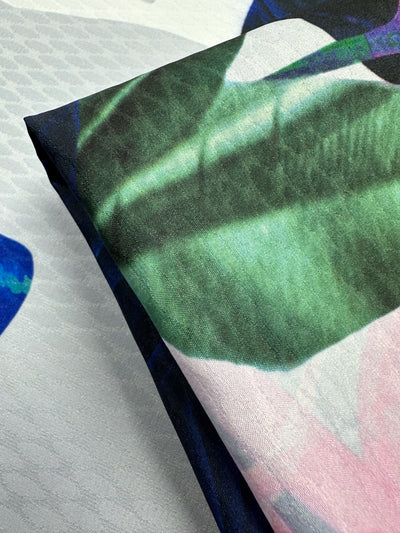 A close-up of Super Cheap Fabrics' "Textured Poly Sateen - Fawna - 150cm" features a folded lightweight fabric with a floral and leaf pattern, showcasing large green leaves accented with pink and blue, ideal for home décor on a light textured background.